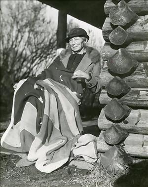 An elderly woman in a hat and winter coat sits in a rocking chair on the porch of a log cabin. A large flag is draped across her lap. 